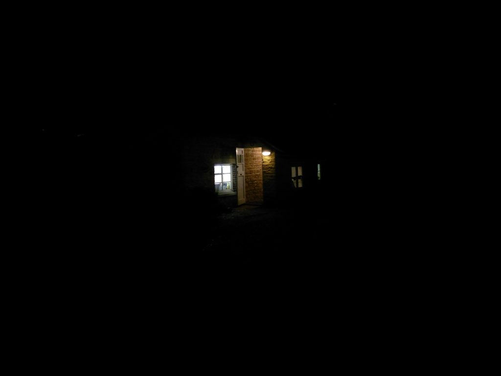 A cottage in pitch black surroundings. From the lights, you can just make out the front door, glowing invitingly.